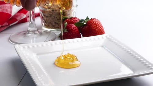 Pouring sweet clover honey unto a white plate to rim a cocktail glass with fresh red strawberries in