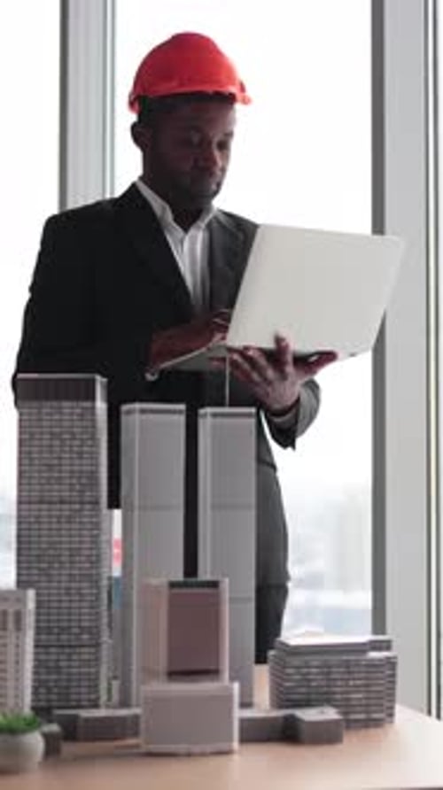 Architect in Hard Hat Using Laptop Inspects Building Model