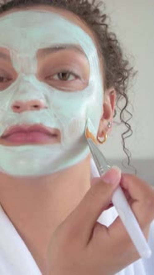 Vidéo verticale : Début de la routine, femme étalant un masque vert avec une brosse dans la chambre à coucher avec un peignoir