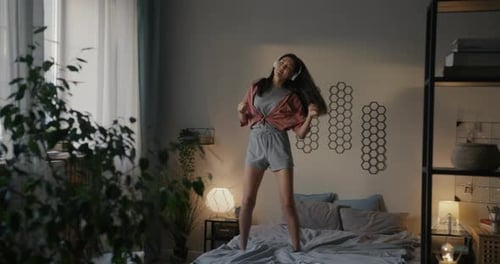 Young Asian Woman Dancing on Bed Wearing Pajamas Listening to Music Enjoying Free Time in Bedroom in