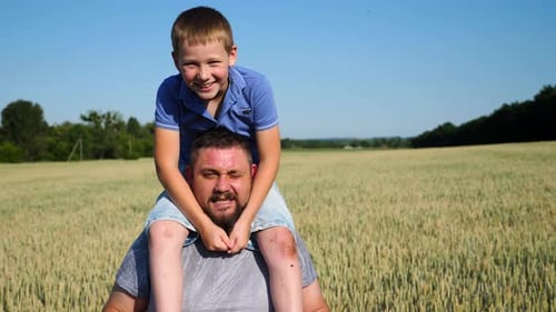 Little Kid Playing with Daddy Beard Sits on His Shoulders Against Background of Cultivated Meadow