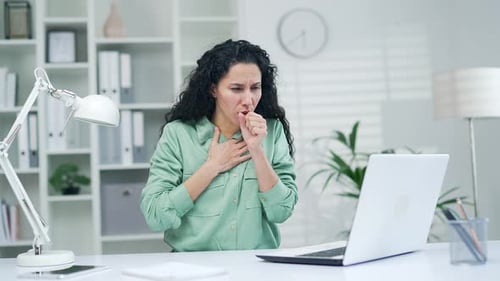 Kranke junge Mitarbeiterin hustet und hält ihre Brust fest. Büroangestellte mit Grippe und Erkältung sitzt a