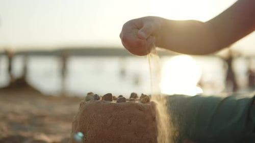 Child Pours Sand on Sand Castle on Shore of Lake at Sunset Closeup