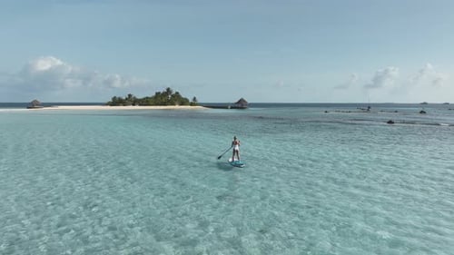Fit woman on standup paddleboard rowing towards tropical island in Maldives