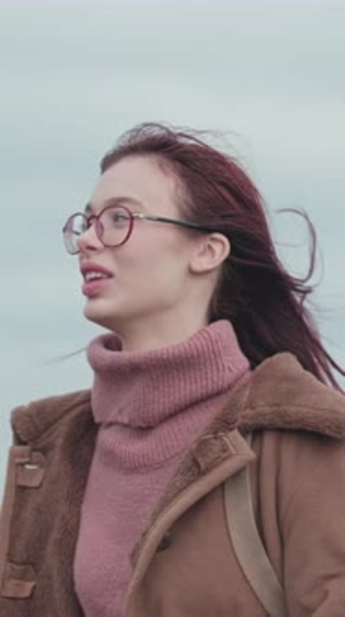 Teenage girl in pink sweater and brown coat giving thumbs up gesture while standing outdoors.