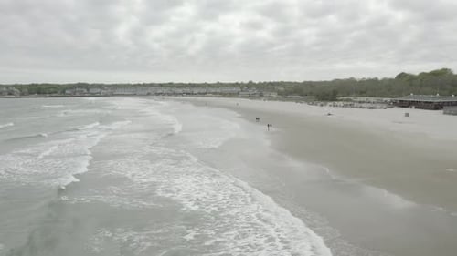 Drone flying to the right and panning to the left, while people enjoy a walk on the beach, cloudy we