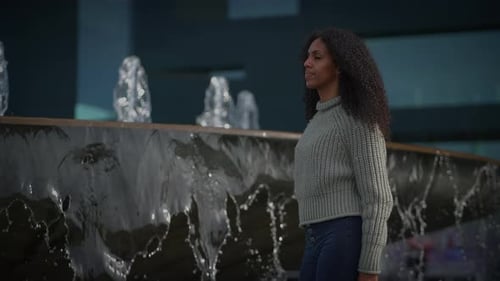 A Young Woman in a Sweater Walks By a Water Fountain at Night Creating a Peaceful Atmosphere
