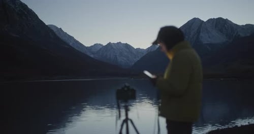 Photographer with Camera and Smartphone By Mountain Lake at Dusk