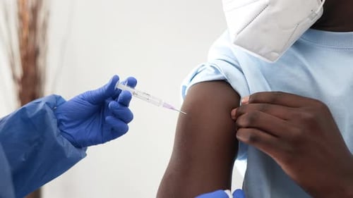 Close Up Of Nurse In Blue Gloves Giving Covid Vaccine To African American Male Patient Wearing Mask