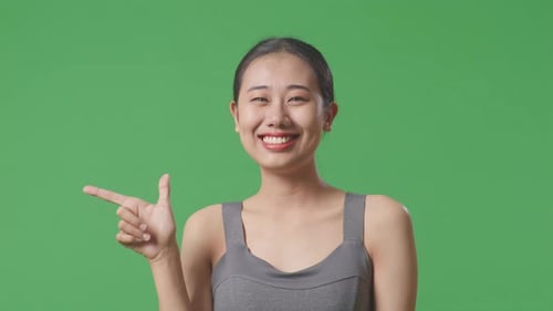 Nahaufnahme einer asiatischen Frau, die Sportkleidung trägt und lächelt, auf grünem Hintergrund zur Seite zeigt