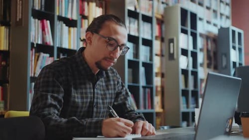 Student Looking at Laptop and Taking Notes in Library