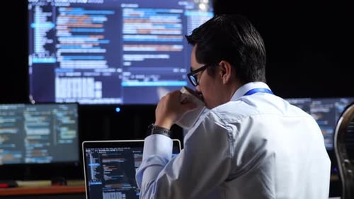 Back View Of Programmer Drinking Coffee While Writing Code By A Laptop Using Multiple Monitors
