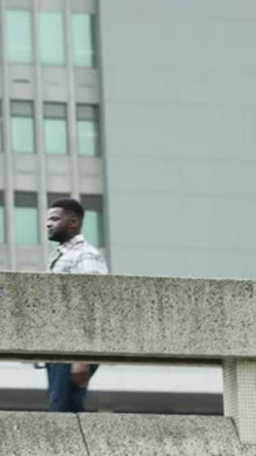 Vertical video: Entering left, man walking office path in plaid shirt, passing railing, foliage