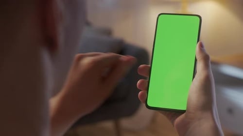 Rückansicht eines Mannes im Wohnzimmer bei Nacht mit Telefon mit grünem Mockup-Bildschirm Chroma Key ohne