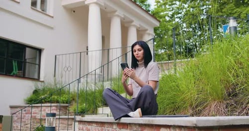 Young Caucasian woman use mobile phone while sitting on the bench near modern old building in the hi