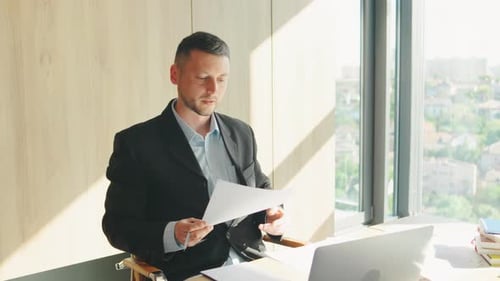 A Focused Businessman is Working in a Modern Office with a City View Carefully Reviewing Important