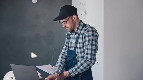 A Construction Engineer at a Construction Site Works with Projects Using a Laptop and Blueprints A