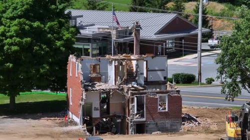 Partially demolished brick house with rubble surrounds. Excavator equipment nearby in a suburban are