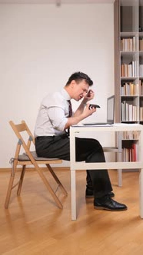 Vertical Video Man with Bad Posture Sitting at a Simple Desk Hunching Over a Laptop While Holding a