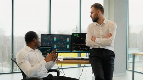 Zwei männliche Händler im Büro überwachen gemeinsam Aktien, Daten, Kerzendiagramme auf dem Bildschirm
