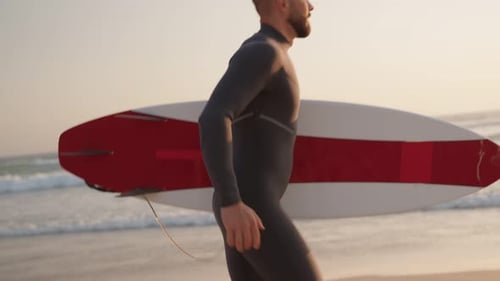 Surfer Walks Along Beach at Sunset with Surfboard Ready to Ride the Waves