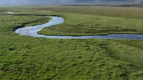 Winding river peacefully meanders through lush green fields. Mountains in the distance. Aerial