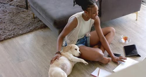 Junge Afroamerikanerin sitzt auf dem Boden, streichelt Hund, benutzt Laptop, zu Hause