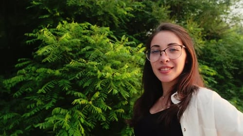 Beautiful girl in green shorts and a white shirt poses on the river bank near a green bush. Looking