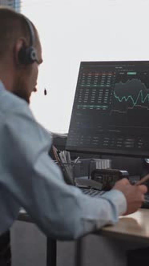 Male Financial Trader in Headset Works on Computer Monitors Stocks Takes Notes