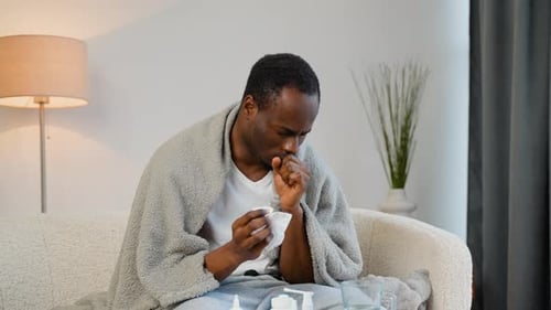 African Man with Runny Nose Sits on the Couch at Home Under a Plaid Sneezing in a Napkin