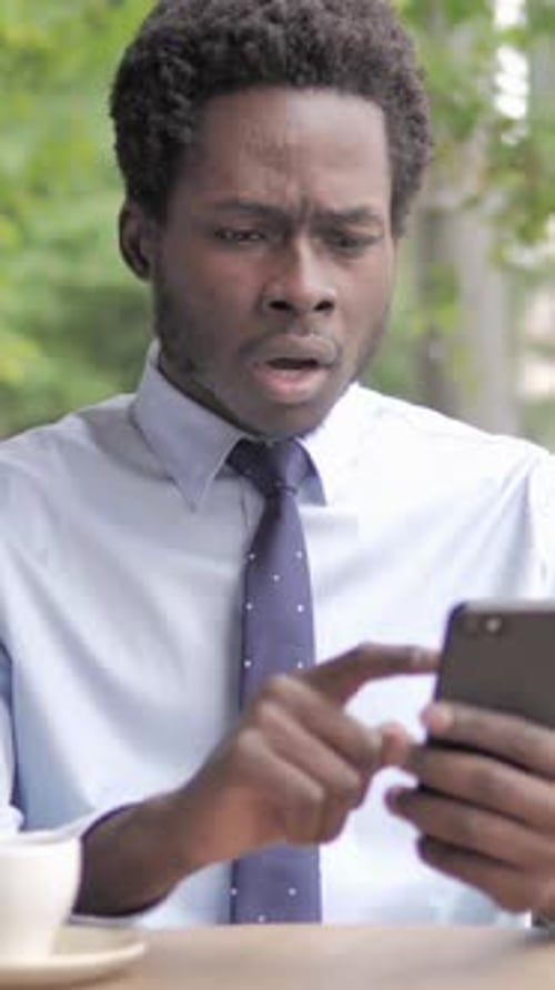 African Businessman Upset by Loss on Smartphone, Sitting in Outdoor Cafe, Vertical Video