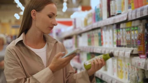 A Woman in a Tan Shirt Stands in a Supermarket Aisle and Scans the Barcode of a Product on the Shelf