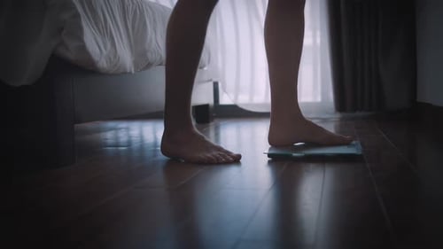 Woman Steps on Scale in Dark Bedroom