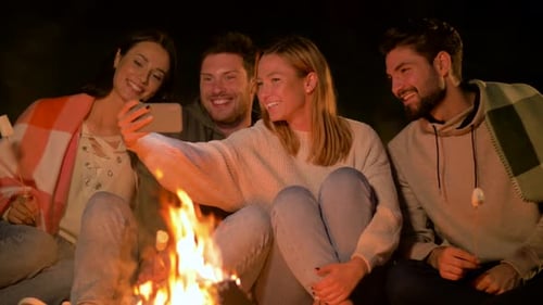 Friends Gather Around Campfire, Look at Phone, at Night