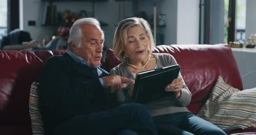 A happy mature couple is using a tablet for family entertainment while sitting on a sofa in living