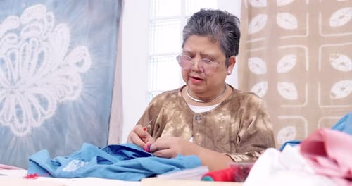 Senior Asian woman artisan pauses, checks red thread end, reviews shibori motif plan, then resumes