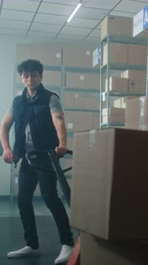 Male Inventory Specialist Pushes Pallet Truck with Cardboard Boxes in Warehouse Facility