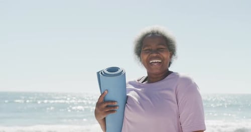 Portrait of happy senior african american woman holding yoga mat at beach, in slow motion