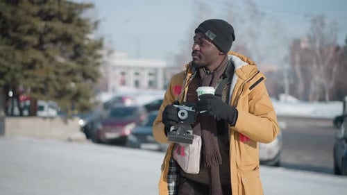 Portraitist Sipping Beverage in Winter City Holding Camera Under Bright Sun