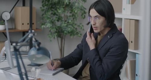 Asian Businessman Speaking on Mobile Phone and Writing in Notebook Working in Office