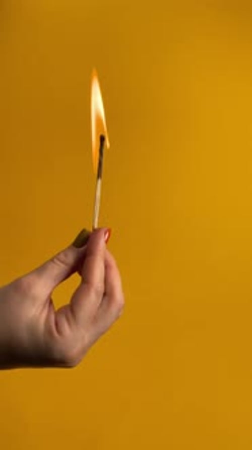 Hand Holding a Burning Matchstick Against Yellow Backdrop