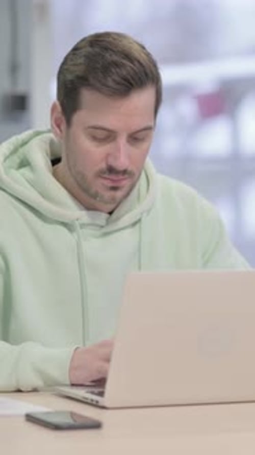Man Working on Laptop Indoors, Wearing a Hoodie