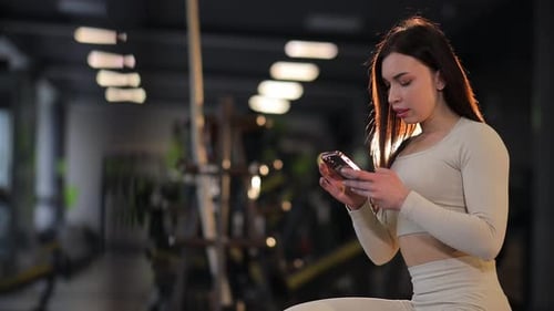 Young Attractive Woman with a Beautiful Figure Looks at a Phone in the Gym