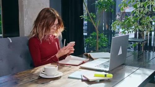 Happy Young Woman Sits with Phone and Computer at the Table