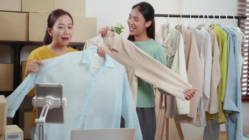 Two young asian woman live online clothing store while showing clothing with tablet on social.