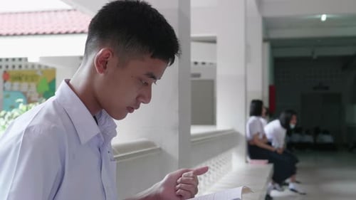 A male Asian high school student in white uniform sitting and reading a book