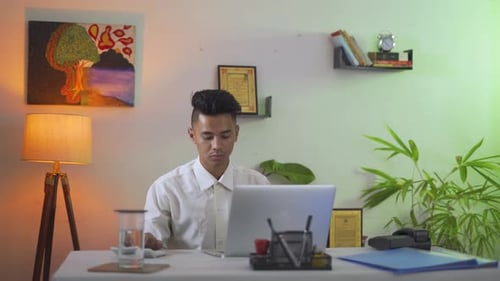 Serious young man working hard in office with his modern laptop device taking a break for drinking h