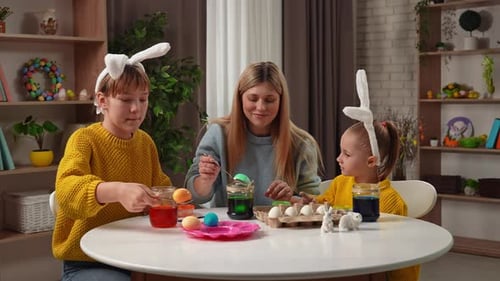 Mother and Little Daughters Preparing for Easter Celebrations Sitting at Table Making Colored Eggs