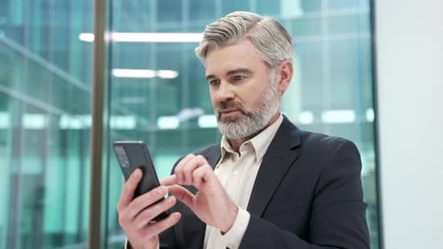 Confident mature businessman is using browsing mobile phone at workplace in business office.