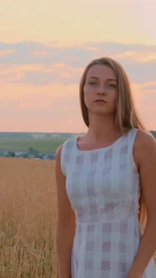 Young Woman in White Dress Standing in Golden Wheat Field During Sunset with Soft Clouds